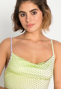 Top sem mangas verde claro com bolinhas roxas, apresentando alças finas e um decote drapeado, feito de um material liso e elástico.