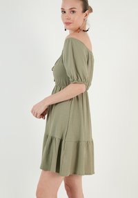 Off-the-shoulder, short-sleeve dress in textured olive green fabric, featuring a cinched waist and tiered ruffle hem.