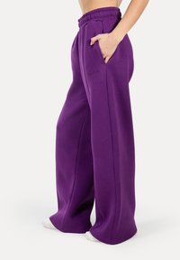 Fialové široké tepláky s elastickým pasem na stahování, bočními kapsami a decentním logem na přední straně. Měkký textura tkaniny.