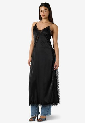 Femme portant une robe nuisette en satin noir avec garniture en dentelle sur un jean bleu et des talons gris ouverts à l’avant, debout sur un fond blanc uni.