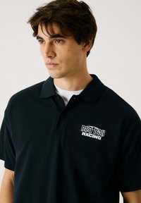 Zwarte poloshirt met een wit "Red Bull Racing" logo op de linkerborst; korte mouwen; zachte katoenstof met een klassieke kraagontwerp.