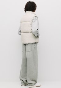 Light beige puffer vest with a high collar, paired with a gray sweatshirt and loose-fitting light gray jeans, and white sneakers.
