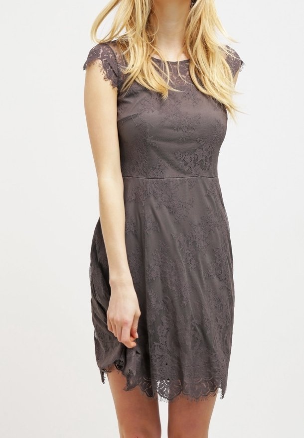 Femme portant une robe en dentelle gris foncé à manches courtes avec un ourlet festonné, debout devant un fond clair uni.
