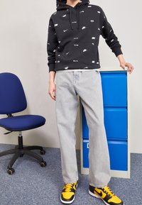 Sudadera con capucha negra con logotipos de Nike en blanco, jeans rectos de pierna en gris claro y zapatillas negras con detalles en amarillo. En el fondo, se observa un archivador azul y una silla.