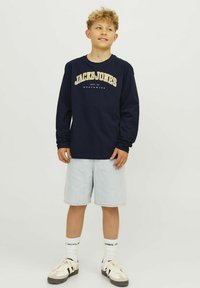 Navy plava majica dugih rukava s žutim grafičkim printom "Jack & Jones", uparena s svijetlim traper kratkim hlačama i bijelim čarapama, s jednostavnim dizajnom tenisica.