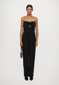 Zwarte jumpsuit met een geplooid lijfje en uitsnijding, met dunne banden en een rechtpijpenontwerp. Inclusief een kleine zwarte handtas.