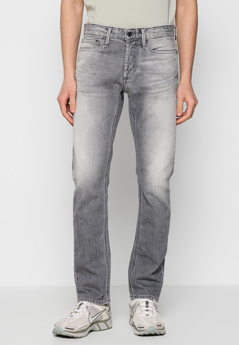 Man in vervaagde grijze slim-fit jeans en witte Nike sneakers, staand tegen een effen witte achtergrond.