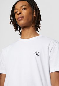 T-shirt bianco in cotone con collo girocollo, dotato di un piccolo logo ricamato "cK" nero sul lato sinistro del petto. Tessuto morbido, vestibilità comoda.