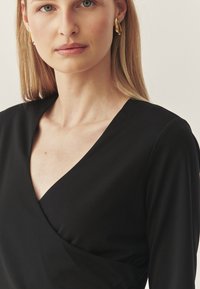 Zwarte wikkelshirt met V-hals, gladde textuur en aansluitend ontwerp. Gouden oorringen accentueren de halslijn.