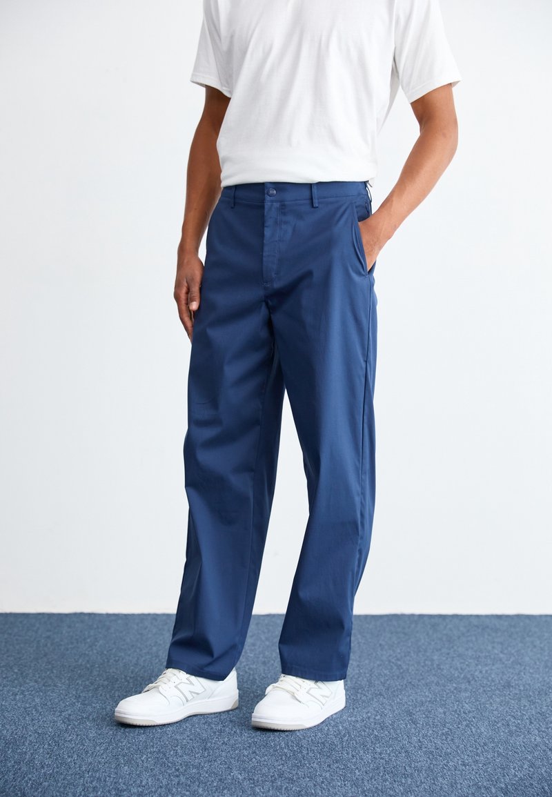 Homme portant un pantalon droit bleu marine, des baskets blanches et une chemise blanche à manches courtes, debout avec une main dans la poche.