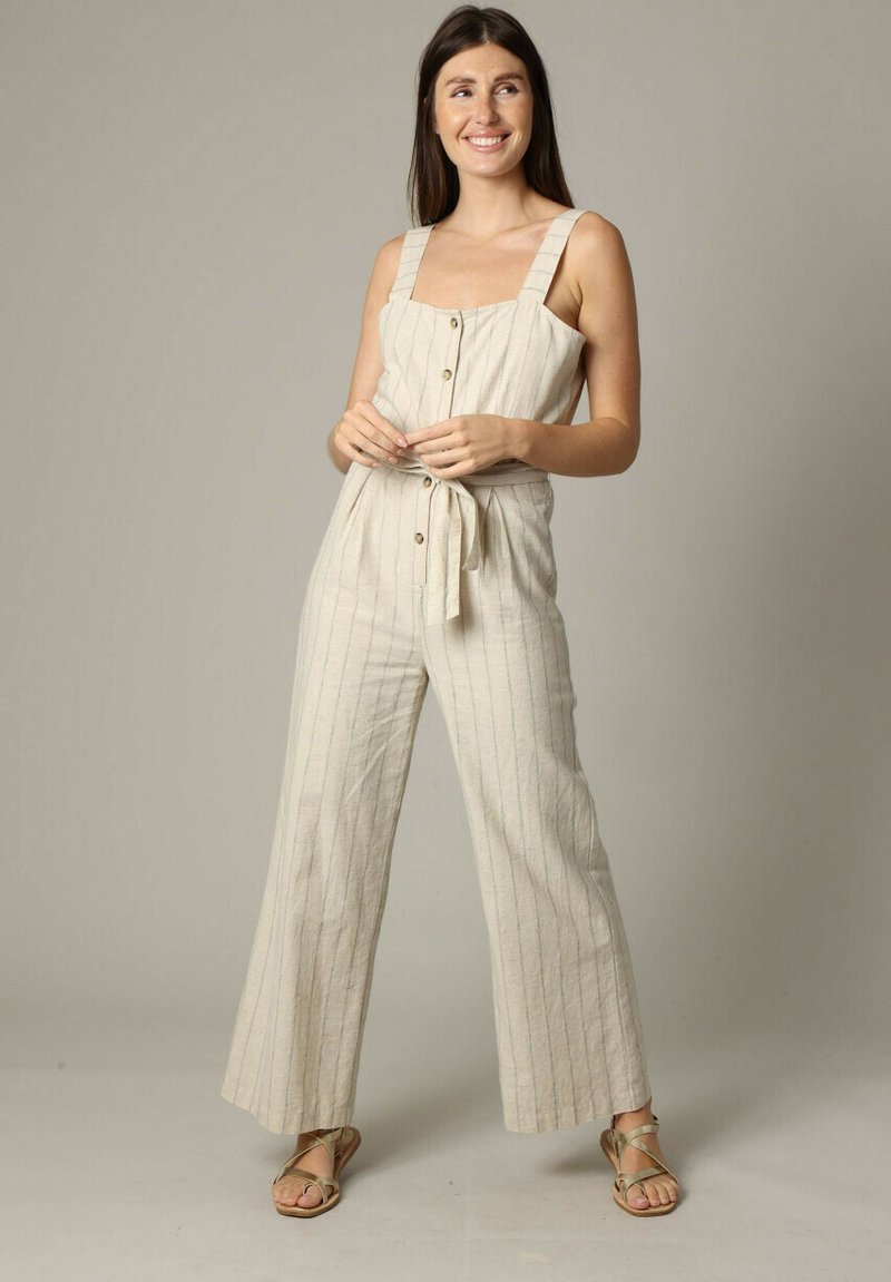 Pièce en lin beige clair avec des rayures verticales, décolleté carré, bretelles larges, fermeture à boutons à l'avant et taille nouée. Des sandales plates complètent la tenue.