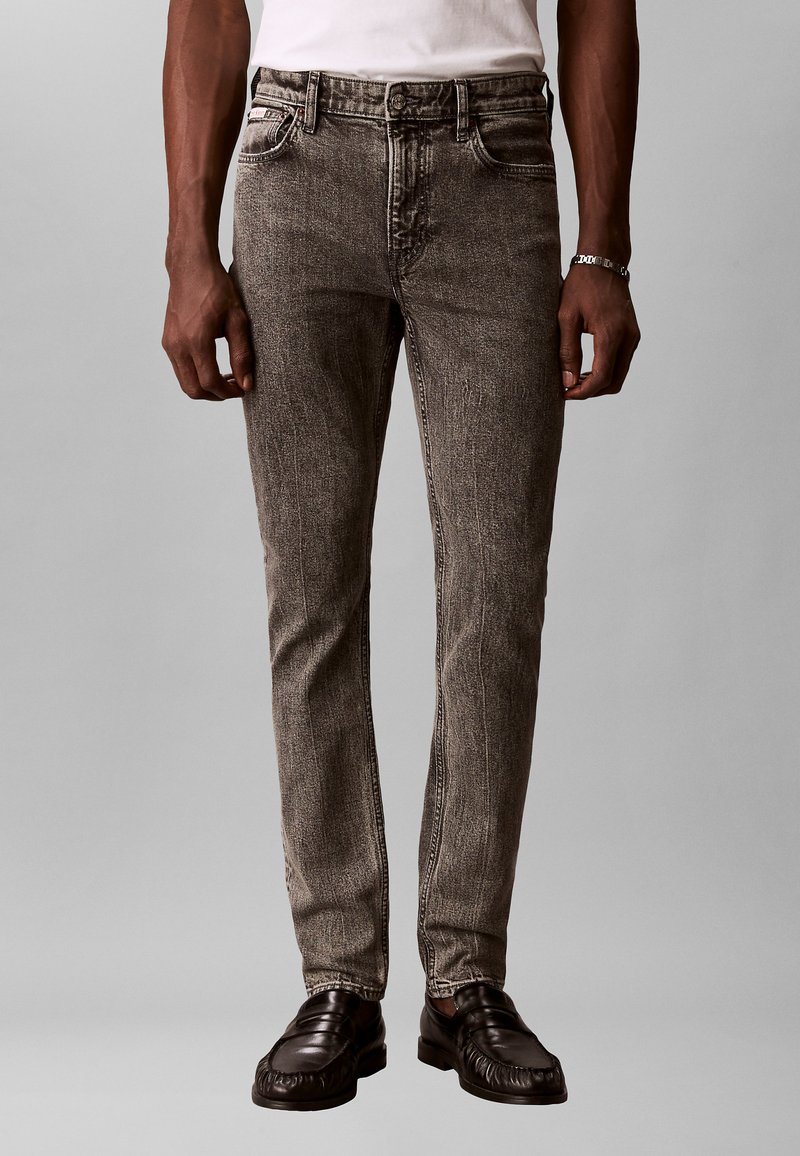 Mann iført smale gråvaskede jeans, svarte skinnloafers, hvit t-skjorte og sølvarmbånd, stående mot en nøytral bakgrunn.