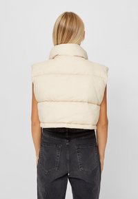 Cream puffer vest with a high collar, sleeveless design, and horizontal quilted pattern, paired with high-waisted black jeans.