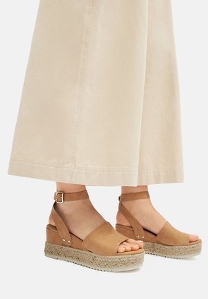 Sandali con zeppa marrone con tomaia in pelle liscia, cinturino regolabile alla caviglia e plateau rivestito in juta. Suola testurizzata per una migliore aderenza.