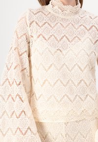 Top in pizzo con intricati motivi a diamante color crema, con alto collo e maniche ampie, che offre un aspetto delicato e testurizzato.