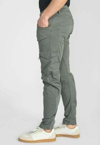 Groene cargo broek met meerdere zakken, ritssluitingen en een taps toelopende pasvorm, gecombineerd met witte sneakers met beige zolen.