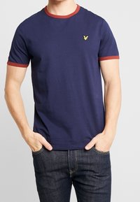 Mężczyzna ubrany w granatową koszulkę z miedzianym wykończeniem kołnierzyka i rękawów, oraz ciemnoniebieskie dżinsy, z rękami w kieszeniach.