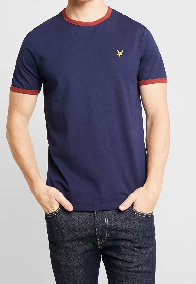 Mężczyzna ubrany w granatową koszulkę z miedzianym wykończeniem kołnierzyka i rękawów, oraz ciemnoniebieskie dżinsy, z rękami w kieszeniach.