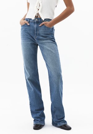 Kvinde iført blå jeans med høj talje og lige ben, hvid cropped ærmeløs top med flæser, hænder i lommerne og sorte sko med firkantet tå.