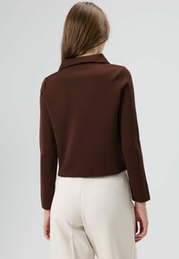 Bruine, gestructureerde stoffen jas met kraag en lange mouwen, iets boven de taille gesneden, gecombineerd met witte, op maat gemaakte broek.