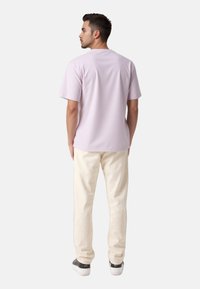 Hellpurpurfarbenes T-Shirt mit kurzen Ärmeln und entspannter Passform, kombiniert mit cremefarbenen Hosen. Von hinten gezeigt, um das schlichte Design und Material zu präsentieren.