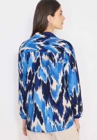 Femme aux cheveux foncés portant une chemise à manches longues avec un motif abstrait bleu, marine et beige, vue de dos sur un fond blanc.