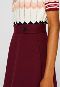 Bordeaux textuurrrok met een aansluitende taille en een zwart knoopdetail. Gepaard met een gemusterde crèmekleurige en roze top met korte mouwen.