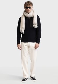 Pull en tricot marine avec une poche, écharpe beige avec des franges, pantalons crème, et baskets noires avec des semelles blanches.