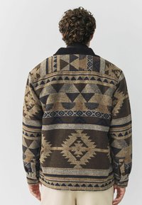 Uomo con capelli ricci che indossa un giubbino a motivo geometrico marrone e beige, in piedi con la schiena rivolta verso la macchina fotografica su uno sfondo semplice.