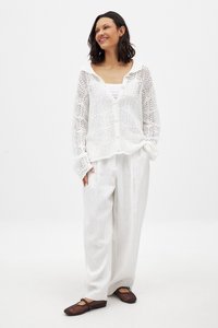 Weißer gestrickter Cardigan mit Knopfverschluss, der ein offenes Webmuster aufweist. Kombiniert mit lockeren weißen Hosen und dunklen, geschlossenen Schuhen.