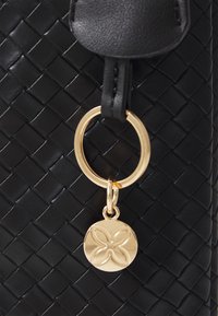 Detalle de un bolso de cuero negro tejido que presenta un anillo de metal dorado con un charm más pequeño que muestra un emblema en forma de flor. Acabado suave y texturizado.