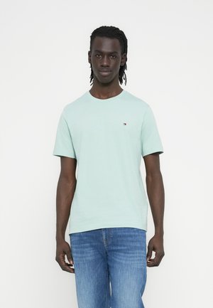 Mann mit dunkler Haut und geflochtenem Haar, der ein hellblaues T-Shirt mit kurzen Ärmeln und blaue Jeans trägt, steht vor weißem Hintergrund.