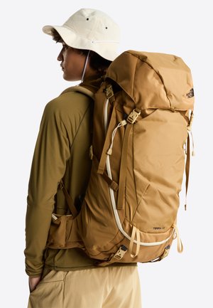 Bruine rugzak voor wandelen met een afgeronde bovenkant, meerdere banden en gespen. Gemaakt van gestructureerde stof. Gecombineerd met een groen shirt met lange mouwen.