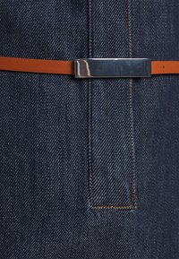 Gros plan sur une ceinture en cuir marron avec une boucle rectangulaire argentée brillante, portée sur un tissu en denim bleu foncé avec des coutures orange.