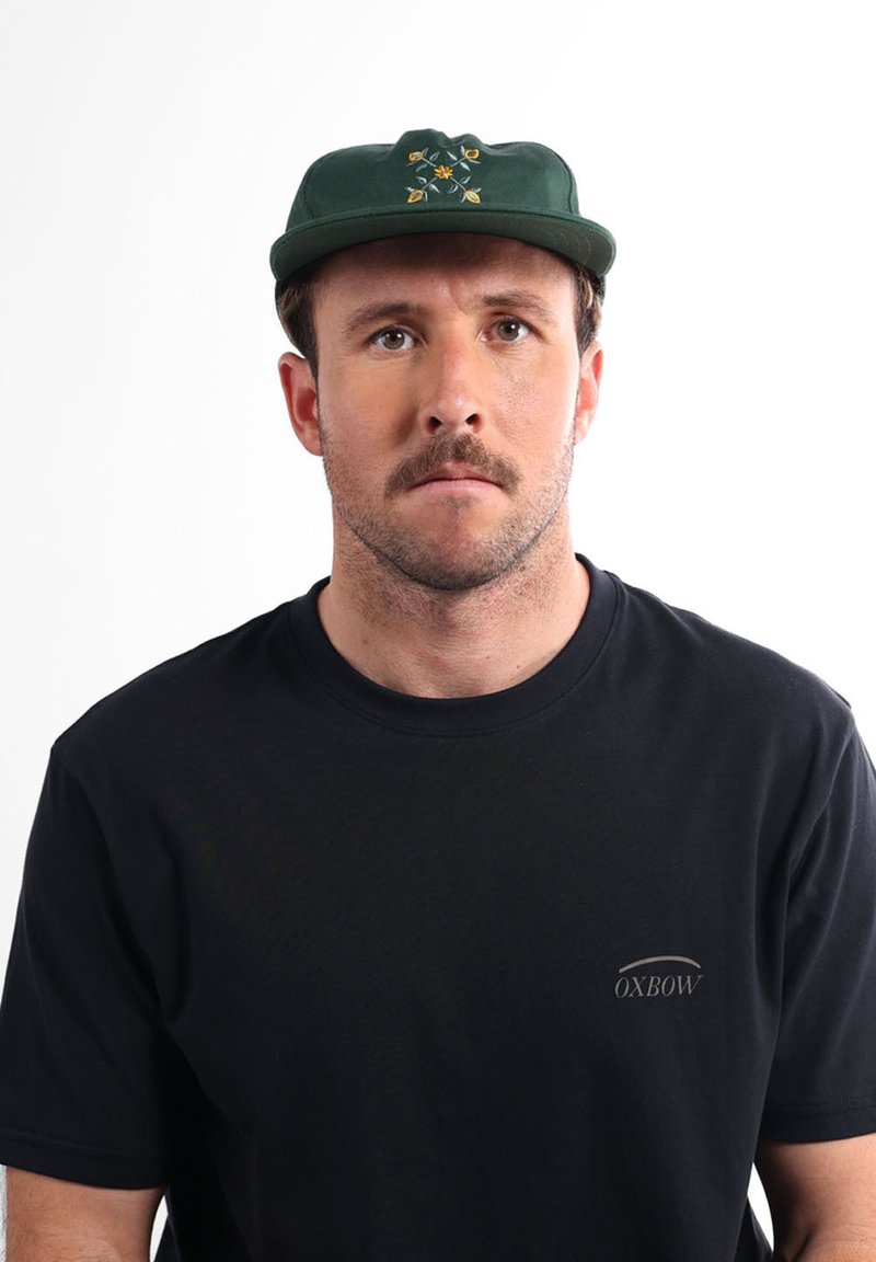 Homme aux cheveux bruns courts et à la moustache, portant une casquette verte à motif floral et un t-shirt noir Oxbow, regardant de face avec une expression neutre.