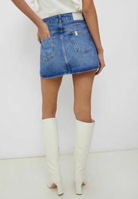 Widok z tyłu osoby w niebieskiej dżinsowej mini spódnicy, białej bluzce i białych wysokich botkach, stojącej na tle gładkiej białej ściany.