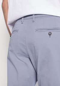 Ljuslila byxor med en slät textur, som har två bakfickor och en synlig knappdetalj. Bärs med en vit t-shirt.