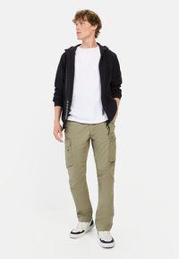 Schwarze Kapuzenjacke mit Reißverschluss, weißes T-Shirt, helle olivfarbene Cargo-Hosen mit Taschen und weiße Sneakers mit dunkelblauen Akzenten. Glatter Stoff, lockere Passform.