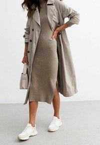 Cappotto trench beige sopra un abito midi rigato verde oliva con uno spacco laterale, abbinato a sneaker bianche e con una piccola borsa bianca.