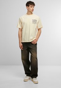 Beige bomulds T-shirt med grafisk tekst på venstre bryst; parret med mørke denim jeans og lyse sneakers med en kraftig sål.