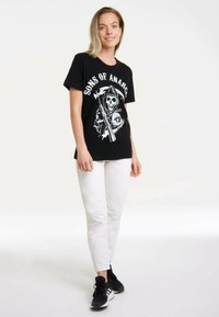 Black cotton t-shirt featuring a white graphic design of a skull and a scythe, paired with white pants and black sneakers.