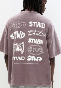 T-shirt grigio oversize con diversi loghi bianchi sul retro, tra cui "STWD" in stili e orientamenti diversi.