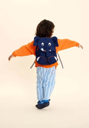 Child with curly hair wears orange sweater, blue striped pants, and navy blue elephant-shaped backpack, arms outstretched, facing away.