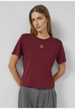 Burgunderfarbiges Baumwoll-T-Shirt mit kurzen Ärmeln, rundem Halsausschnitt und einem kleinen dekorativen Detail auf der Brust. Kombiniert mit grauen Nadelstreifenhosen.