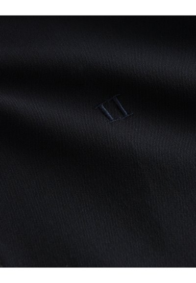 Tissu noir avec un discret chiffre romain II brodé en fil noir assorti, montrant une texture fine et de légers plis.