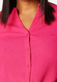 Chemise boutonnée rose vif avec un tissu texturé. Dotée d'un col en V et de boutons visibles le long de la patte de boutonnage avant.