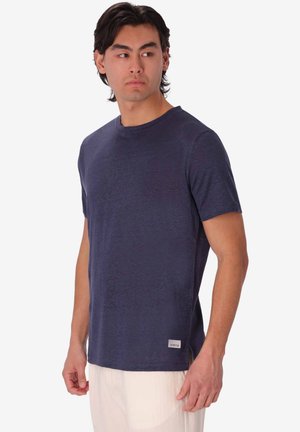 Uomo che indossa una maglietta blu navy a maniche corte e pantaloni chiari, guarda di lato su uno sfondo neutro.