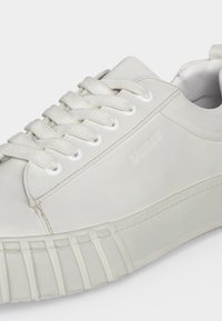 Λευκό παπούτσι τύπου sneaker με χαμηλή γλώσσα, υφασμένες κορδόνια, ανάγλυφο όνομα μάρκας στο πλάι και παχύ λάστιχο σόλας με γεωμετρικά σχέδια.