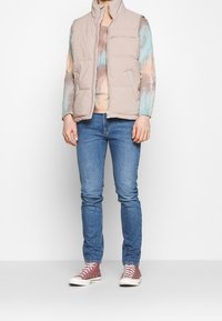 Chaleco acolchado sin mangas beige con cuello alto y bolsillo frontal con cremallera, usado sobre una camisa de manga larga tie-dye y jeans azules.