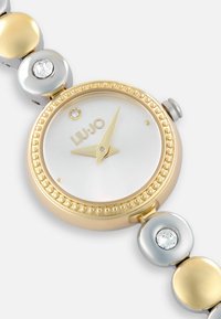 Reloj de pulsera Liu Jo con caja redonda en tono dorado, esfera blanca, eslabones en oro y plata, y un acento de cristal en la correa para un detalle.